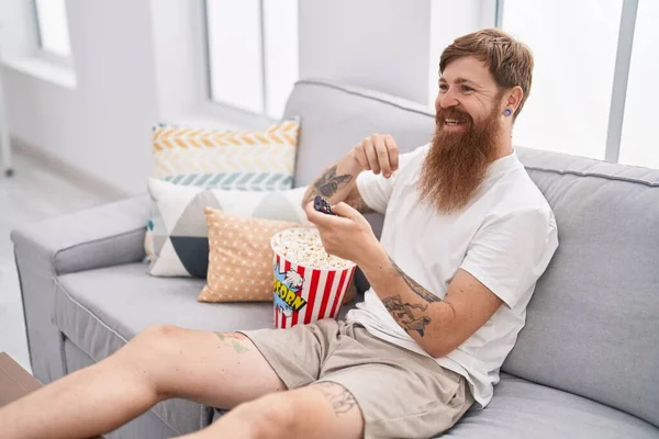 Young redhead man watching movie sitting on sofa at home