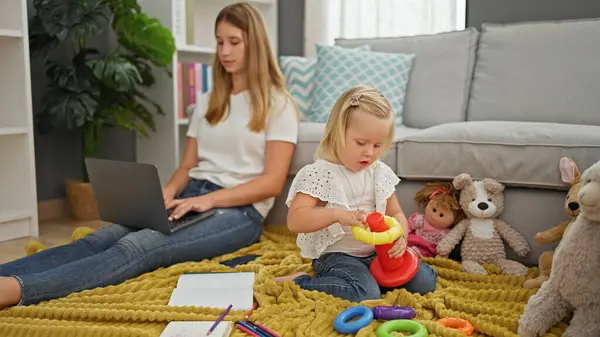 Serious caucasian mother teleworking on laptop, together with relaxed little girl playing with doll on floor blanket in living room at home