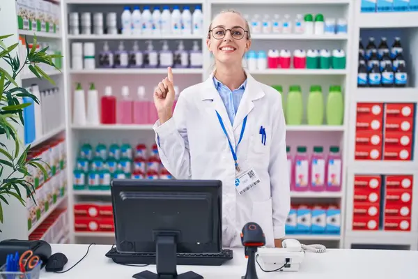 Young caucasian woman working at pharmacy drugstore showing and pointing up with finger number one while smiling confident and happy.
