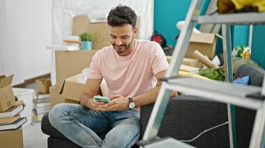 Genç İspanyol adam akıllı telefon kullanıyor. Yeni evindeki kanepede oturuyor.
