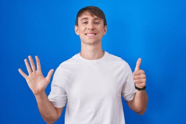 Kafkasyalı sarışın adam mavi arka planda duruyor. Altıncı parmaklarıyla işaret ediyor ve gülümsüyor. Kendinden emin ve mutlu.. 