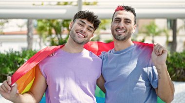 İki erkek parkta homoseksüel bayrağı tutarak birbirlerine sarılıyorlar.