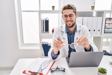 Young man optician holding glasses using laptop at clinic