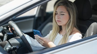 Genç sarışın kadın akıllı telefon kullanıyor. Sokakta arabada oturuyor.