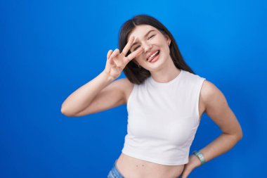 Young caucasian woman standing over blue background doing peace symbol with fingers over face, smiling cheerful showing victory 