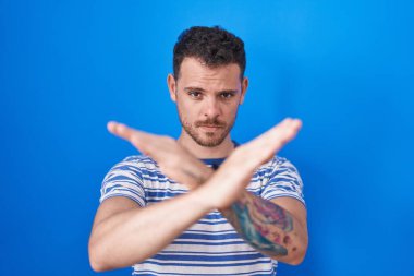 Young hispanic man standing over blue background rejection expression crossing arms doing negative sign, angry face 