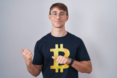 Beyaz, sarışın, bitcoin tişörtlü bir adam. Arka tarafı işaret ediyor. El ve baş parmakları havada, kendinden emin. 