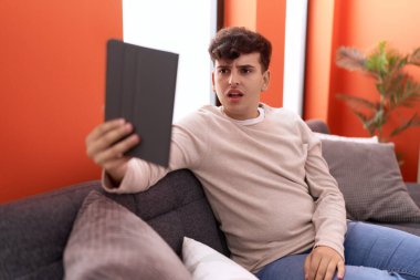 Kanepede oturan, dokunmatik ped kullanan genç, korkmuş ve ağzı açık, şaşırmış, inançsız bir adam. 