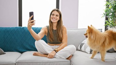 Neşeli, genç İspanyol kadın evinde, oturma odasındaki kanepesinde rahatça gülümsüyor. Evcil köpeğiyle mutlu bir selfie çekmek için akıllı telefon teknolojisi kullanıyor..