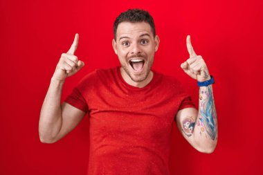 Young hispanic man standing over red background smiling amazed and surprised and pointing up with fingers and raised arms. 