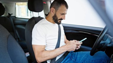 Young hispanic man using smartphone sitting on car at street