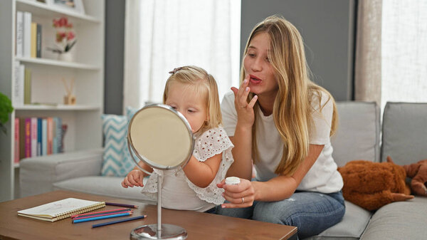 Relaxed caucasian mother and daughter cosy at home, lying comfortably on sofa, seriously applying makeup on lips, looking into mirror