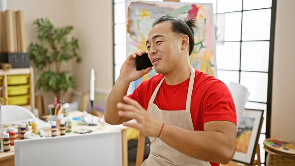 Confident young chinese artist, cheerfully talking on smartphone in his art studio, filled with canvas and vibrant paint palette