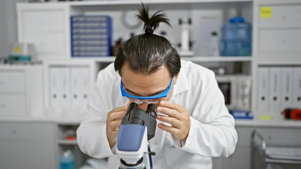 Focused young chinese man, a dedicated scientist at the heart of medical technology, deeply immersed in biology research using microscope in lab.