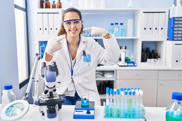 Young hispanic woman working at scientist laboratory smiling cheerful showing and pointing with fingers teeth and mouth. dental health concept.