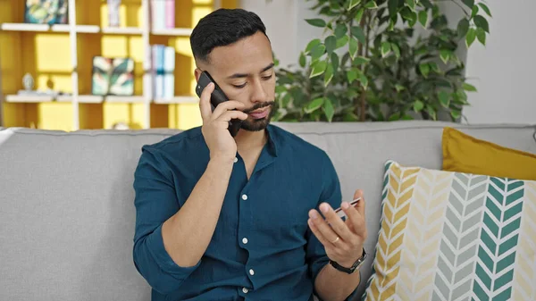 Young hispanic man speaking on the phone holding credit card at home