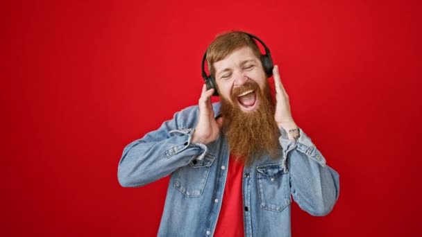 音楽を聴く若い赤毛の男は 孤立した赤い背景の上に親指でジェスチャーを上げる — ストック動画