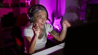 Neşeli, orta yaşlı, gri saçlı kadın flamalı, oyun konsolunu güvenle oynuyor. Oyun sahasında galibiyetini kutluyor. Oyun odasında, gülümseyen bir kulaklıkla kazanıyor.!