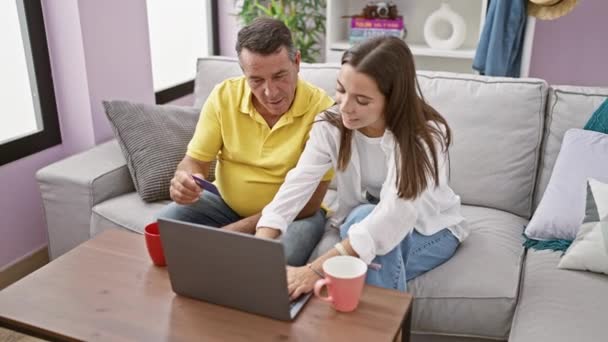 西班牙裔父亲和微笑的女儿在家里网上购物 坐在客厅沙发上 手握笔记本电脑和信用卡 一起喝着一杯浓缩咖啡 — 图库视频影像
