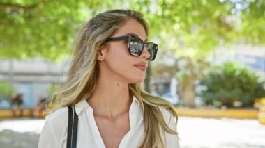 Güzel sarışın kadın, ciddi bir ifadeyle yan tarafa bakıyor. Serin güneş gözlüğünü çıkarıyor, güneşli parkta rahatça duruyor.