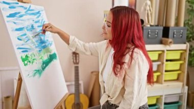 Enfes, kendine güvenen genç kızıl saçlı sanatçı fırçalar ve tuvallerin arasında hareketli bir sanat stüdyosunda neşe içinde çizim yapıyor.