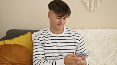 Genç, yakışıklı bir İspanyol 'un akıllı telefonunu neşeyle kullanması. Gülümsüyor, kanepesinde oturuyor, mutlu mesajlar atıyor. Mesajları neşeli bir şekilde yazmak, kapalı mekanda teknoloji zamanının tadını çıkarmak..