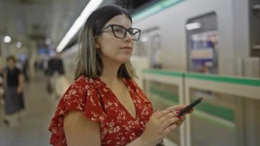 Göz kamaştırıcı İspanyol kadın, gözlüklü, yeraltı tren istasyonunda duruyor, metro yolculuğunu tahmin ediyor, telefonuna dalmış.