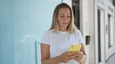 Şık, genç, sarışın bir kadın akıllı telefonuyla online mesaj göndermeye odaklanmış, güneşli şehir caddesinde duruyor, havalı bir şehir hayatı yaşıyor..