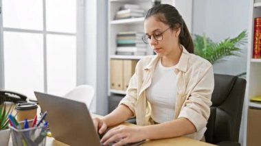 Genç, güzel İspanyol kadın iş sihrini bir dizüstü bilgisayarla yapıyor. Profesyonel bir ofis içinde neşe ve başarı yayıyor..