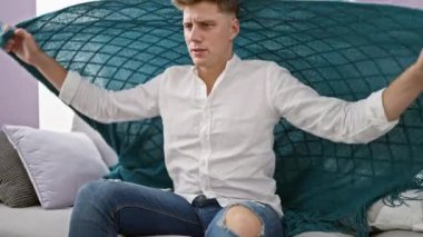 Genç beyaz adam kanepede oturuyor ve evinde üşütmek için battaniyeyi örtüyor.