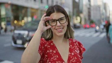 Gülen, kaygısız, güzel gülüşlü ve gözlüklü bir İspanyol kadın, kendinden emin bir şekilde Tokyo şehrinin modern caddesinde poz veriyor, bulaşıcı bir şekilde neşeli ve pozitif bir aura yayıyor.