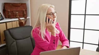 Genç, çekici sarışın kadın, iş konuşmalarıyla ofisteki akıllı telefon ve dizüstü bilgisayarla rahatlıyor, başarılı ve profesyonel bir şekilde ceketine ve gözlüğüne güveniyor.