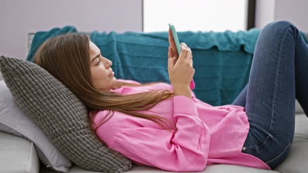 自宅で陽気な若いヒスパニックの女性は ソファーに横たわり スマートフォンで楽しくテキストを送り 喜びとポジティブさを放つ — ストック動画