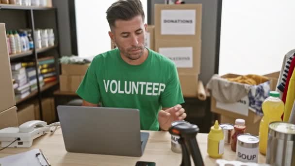 チャリティーセンターで献身的な若いヒスパニック系男性ボランティア ノートパソコンで働いている間に缶詰食品の寄付を処理 — ストック動画