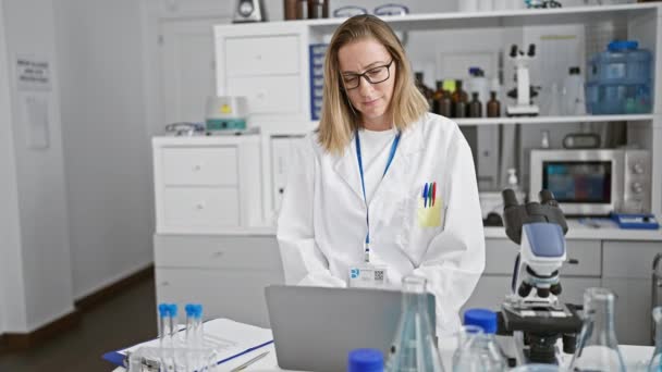 疲惫的金发科学家放松了下来 年轻女人在实验室辛辛苦苦地工作了一天后伸出手来 摘下眼镜 靠在笔记本电脑上 — 图库视频影像