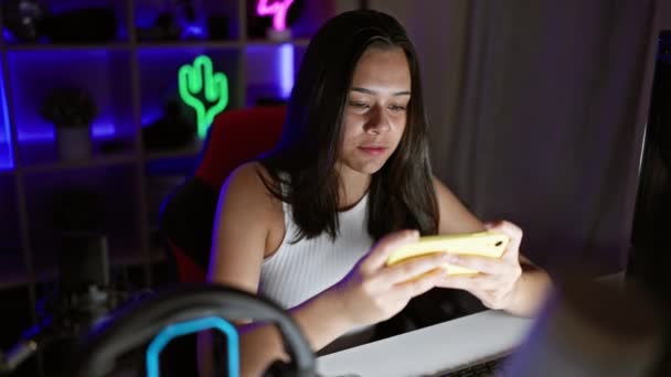 ゴージャスな若いヒスパニック女性がゲームルームでスマートフォンを使用してゲームセッションをストリーミングし 自信を持って微笑みます — ストック動画