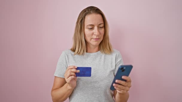 ハッピーで若いブロンドの女性は自信を持ってスマートフォンに支払いメッセージを入力し クレジットカードを保持し 気軽に立っています ピンクの隔離された背景の壁の美しい表現 — ストック動画