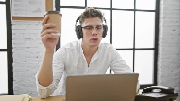 英俊的年轻白种人 一位自信的生意人 在耳机上哼唱着音乐 在执行办公室的办公桌边喝着早晨的咖啡 — 图库视频影像