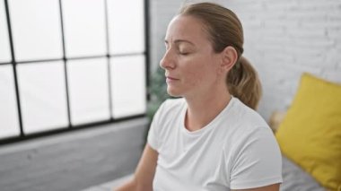 Güzel, ciddi sarışın bir kadın. Yoga egzersizleriyle sakin dengede duruyor. Yatak odasının içinde odaklanmış bir konsantrasyonda oturuyor..