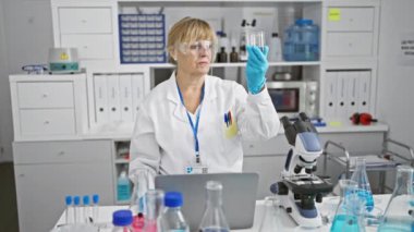 Olgun, sarışın bir bilim adamı, orta yaşlı bir kadın, test tüpündeki sıvıyı titizlikle ölçerken dolup taşan bir laboratuarda laptopuna notlar alıyor..