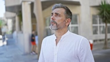 Kendine güvenen, sakallı, genç İspanyol, kır saçlı, şehir merkezindeki bir caddenin ortasında, gökyüzünün altında dikilen, o anda gülümseyen, geç kalma endişesi olmayan,.