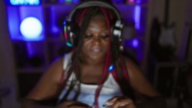 Oyun odasının kalbinde parlayan Afrikalı Amerikalı kadın flaması, gece seansı, dijital eğlencenin ortasında kollarını açmak, kulaklık takan bir oyuncu kraliçesinin güzel portresi.