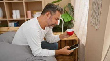 Genç Latin asıllı, kır saçlı, akıllı telefon kullanan, yatakta kahve içen adam.