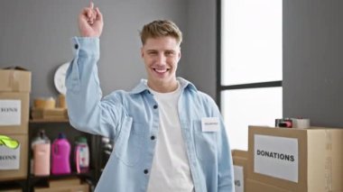 Kendine güveni tam beyaz bir adam, hayır kurumunda gönüllü olarak bağış kutularının arasında dikilirken kameraya bakıyor.