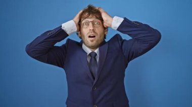 Çılgın stresli genç adam, elleri başını tutuyor, mavi takım elbise içinde şok olmuş ve korkmuş. Şaşırtıcı bir ifade, açık bir ağız, vahşi sürprizin izole edilmiş arka planı. Hüsrana uğramış, deli ama yakışıklı bir yetişkin..