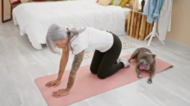 Sportif orta yaşlı kadın, gri saçlı ve formda, sevgili köpeğiyle yoga eğitimi veriyor, yatak odasının rahatlatıcı atmosferinde onu geriyor.