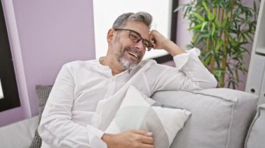 Neşeli, gri saçlı İspanyol adam evdeki oturma odasındaki kanepede mutlu ve rahat bir şekilde oturur, güvenle gülümser..