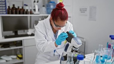 Gülümseyen kızıl saçlı kadın bilim adamı. Laboratuvarda mikroskop kullanıyor. Kolları çapraz oturarak keşif ve analiz yapıyor..