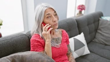 Gri saçlı orta yaşlı kadın, evde köpekle takılıp, kanepede endişe verici telefon konuşmaları yapıyor.
