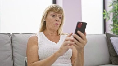 Teknoloji zamanından zevk alan neşeli orta yaşlı sarışın kadın, akıllı telefonu işaret ederken gülümsüyor, evdeki oturma odasındaki kanepede rahatça oturuyor.